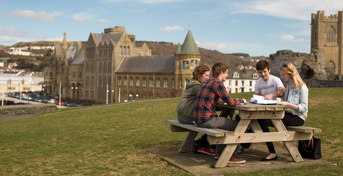 Grŵp o bobl yn eistedd wrth fwrdd picnic yng ngerddi Castell Aberystwyth