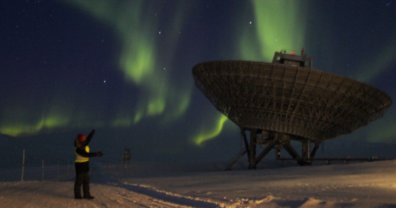 Y myfyrwyr ar y radd MPhys Ffiseg gyda Ffiseg y Planedau a'r Gofod, Elliot Vale yn sefyll ger radar EISCAT ar Svalbard a goleuni’r gogledd yn awyr y nos.