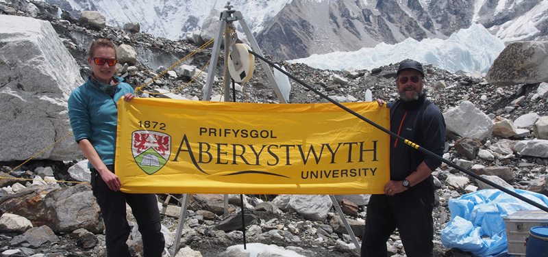 Aelodau Prifysgol Aberystwyth o brosiect EverDrill, Katie Miles a’r Athro Bryn Hubbard, yn chwifio’r faner o flaen safle dyllu rhif 3 ar rewlif Khumbu, nepell o Wersyll Cyntaf Everest.