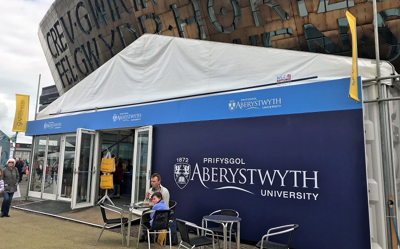 Mae stondin Prifysgol Aberystwyth yn Eisteddfod Genedlaethol yr Urdd eleni reit gyferbyn â Chanolfan y Mileniwm,