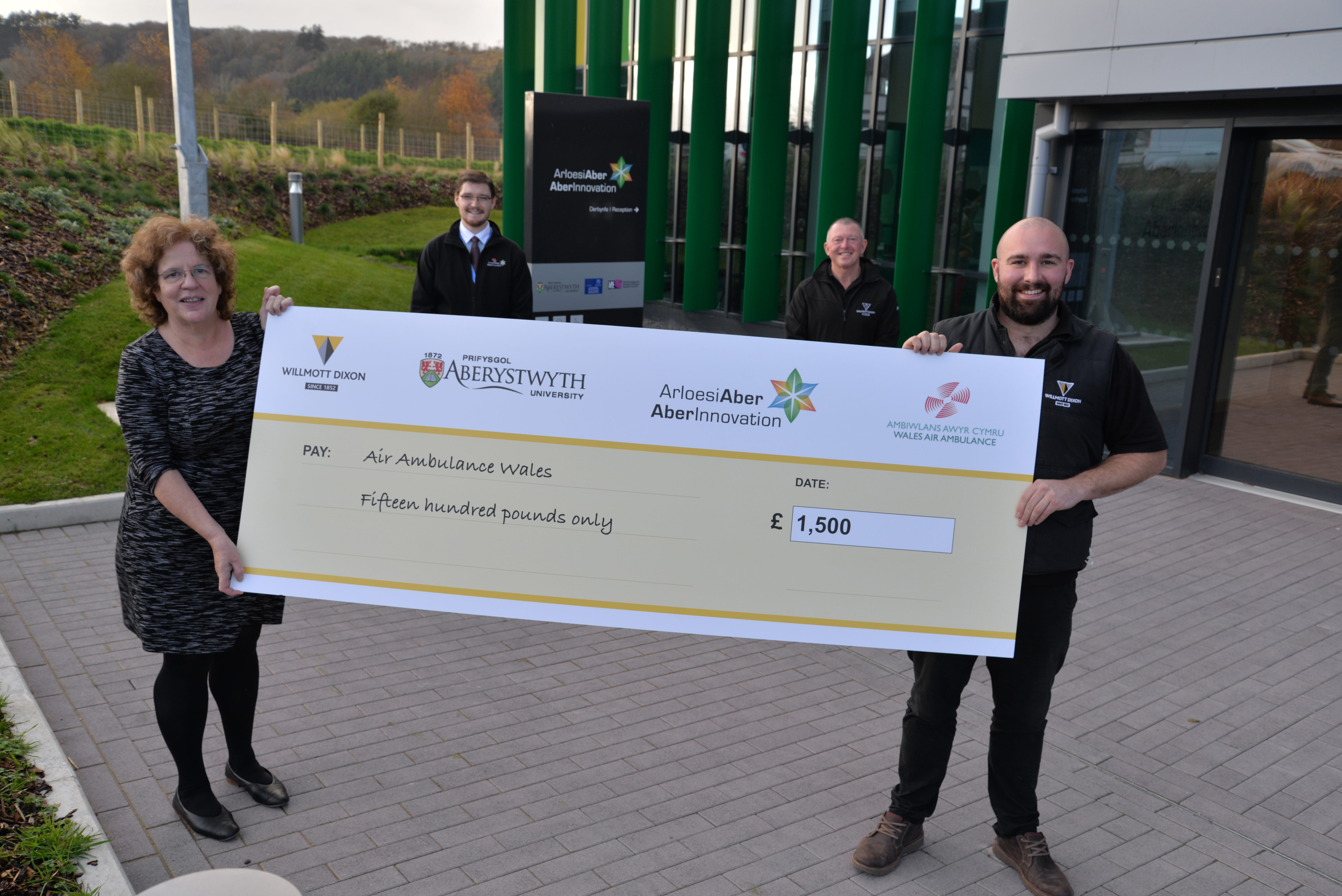 Yr Athro Elizabeth Treasure, Is-Ganghellor Prifysgol Aberystwyth, yn derbyn siec am £1,500 i Ambiwlans Awyr Cymru gan Andrew Jones (Rheolwr Adeiladu Cynorthwyol), Phil Williams (Rheolwr Adeiladu Cynorthwyol) a Shaun Davies (Cynorthwyydd Gweithrediadau) o Willmott Dixon.