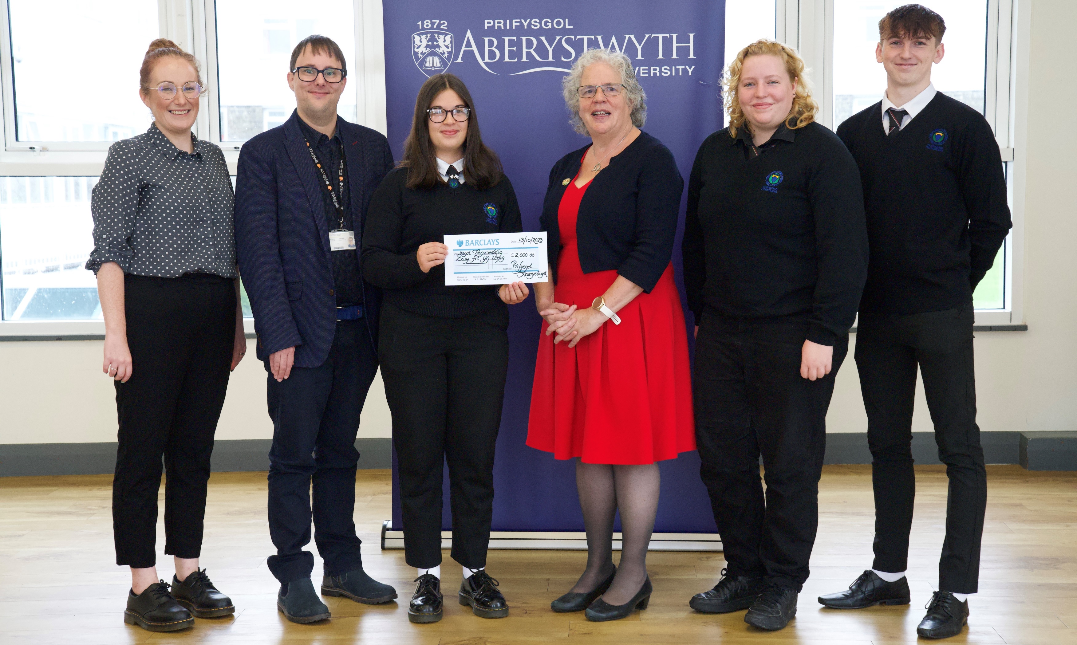 Aelodau o dîm buddugol Ysgol Penweddig Maria Jones, Elain Tanat Morgan (ail o’r dde) a Noa Rowlands yn derbyn siec am £2000 gan yr Athro Elizabeth Treasure, Is-Ganghellor Prifysgol Aberystwyth yng nghwmni Jane Richards, Pennaeth Busnes yn Ysgol Penweddig (chwith pellaf) a Mr Jonathan Fry o Ysgol Busnes Aberystwyth.