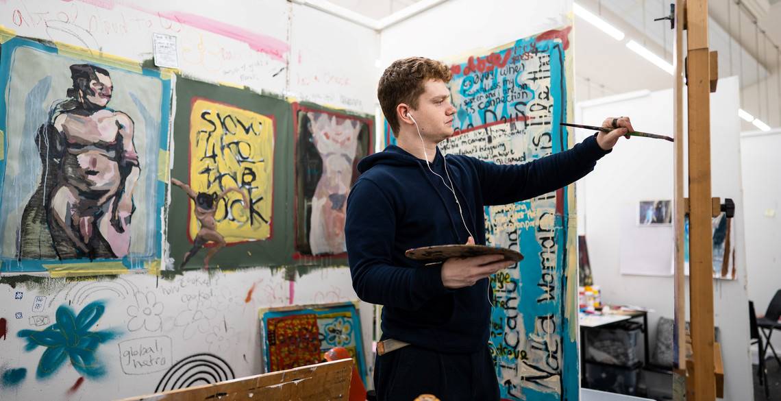 Man painting on an easel surrounded by artwork
